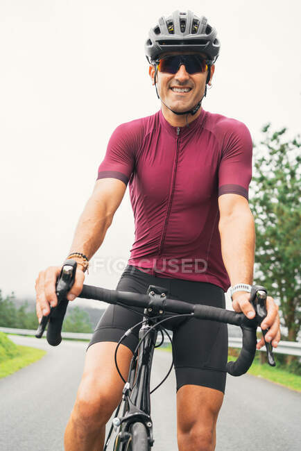 Desportista adulto alegre em óculos de sol de ciclismo e capacete sentado na bicicleta de estrada na estrada rural à luz do dia — Fotografia de Stock