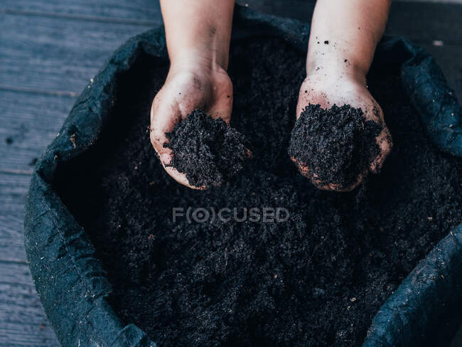 Alto ángulo de cultivo irreconocible niño extendiendo las manos y demostrando tierra seca contra maceta durante el día - foto de stock