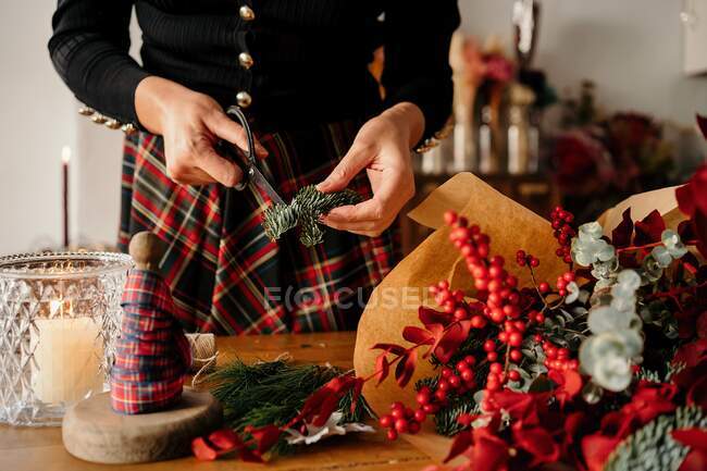 Crop unrecognizable female florist standing and cutting twigs of fir while arranging Christmas bouquet on wooden table — Stock Photo