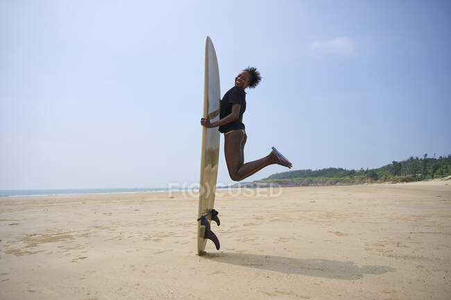 Side view of happy African American female surfer in thong with longboard surf jumping above sandy coast under cloudy blue sky — Stock Photo