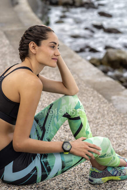 Side view of slim female athlete in sportswear sitting on stone promenade near sea and having break during training — Stock Photo