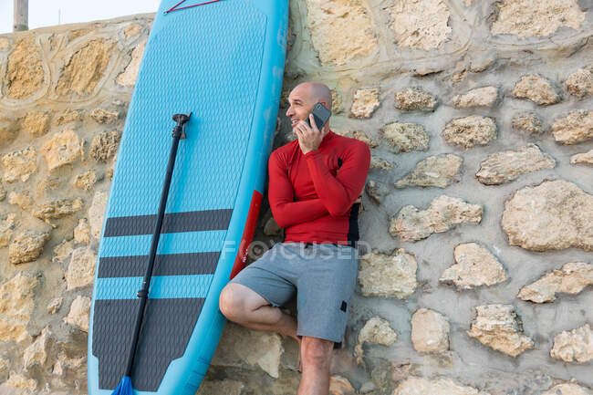 Surfer im Neoprenanzug lehnt an Steinmauer und spricht auf Smartphone mit Paddel und SUP-Brett, während er sich auf das Surfen am Meer vorbereitet — Stockfoto