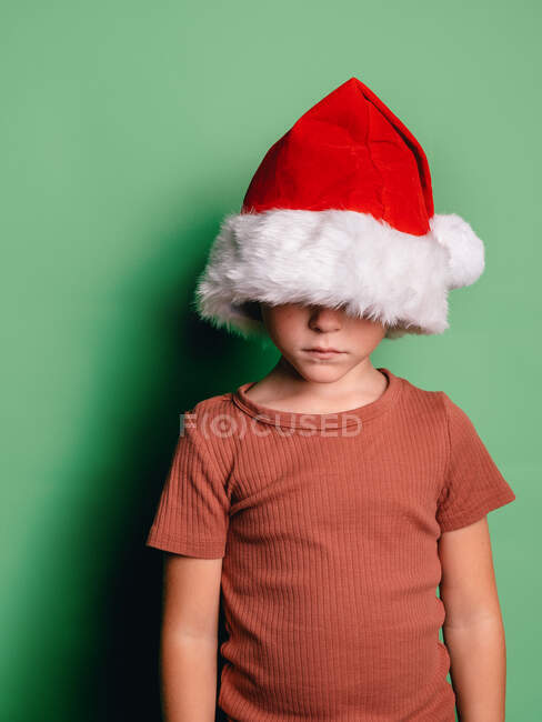 Unrecognizable serious boy covering face with red Santa hat standing against green background — Stock Photo