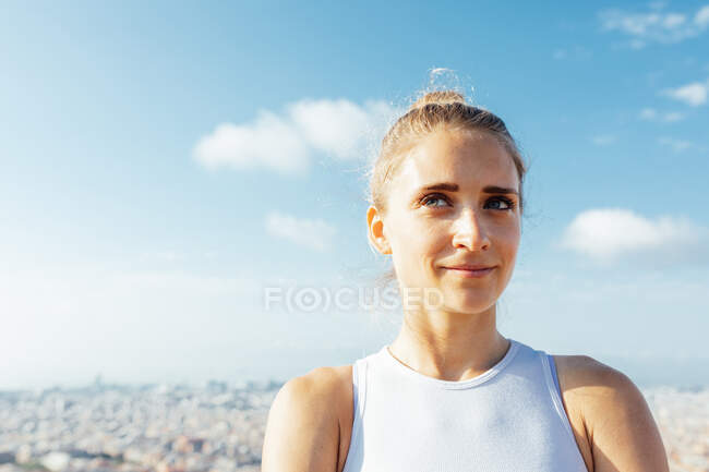Soñadora joven atleta admirando la ciudad mientras mira hacia otro lado bajo el cielo azul nublado a la luz del sol - foto de stock