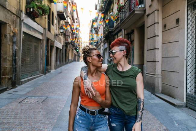 Coole junge homosexuelle Frauen mit Tätowierungen in Sonnenbrillen schauen einander an, während sie sich auf dem Gehweg in der Stadt umarmen — Stockfoto