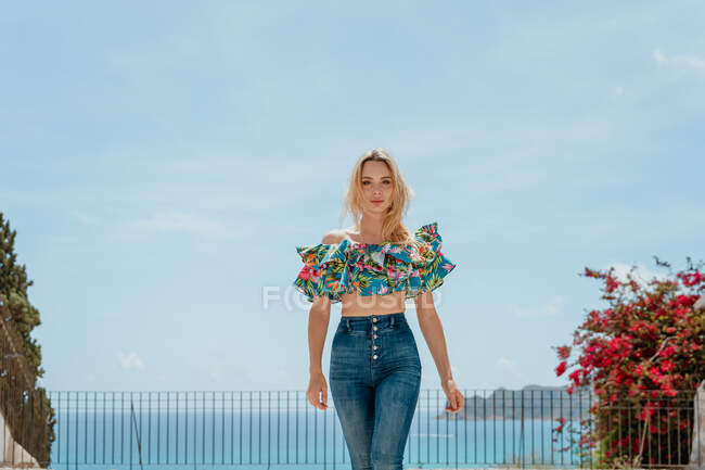 Junge Frau in trendigen Jeans und Top steht auf dem Dach eines Hauses an der Küste des blauen Meeres — Stockfoto