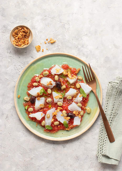 Sommerlicher Tomaten-Kabeljau-Salat in einer flachen Schüssel mit Gabel und einer Schüssel gebratener Zwiebeln — Stockfoto