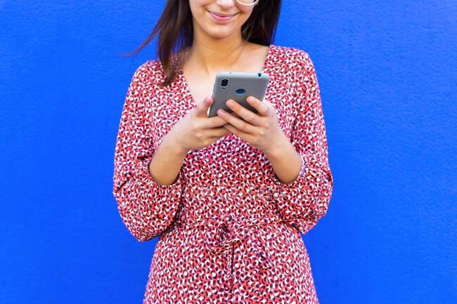 Обрізана невпізнавана усміхнена жінка в сукні та окулярах, що стоїть біля синьої стіни та використовує смартфон удень — стокове фото