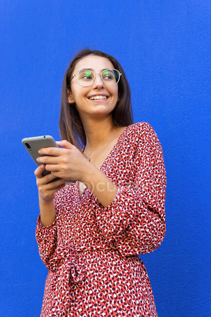 Mulher sorridente em vestido e óculos de pé olhando para longe perto da parede azul e usando smartphone durante o dia — Fotografia de Stock