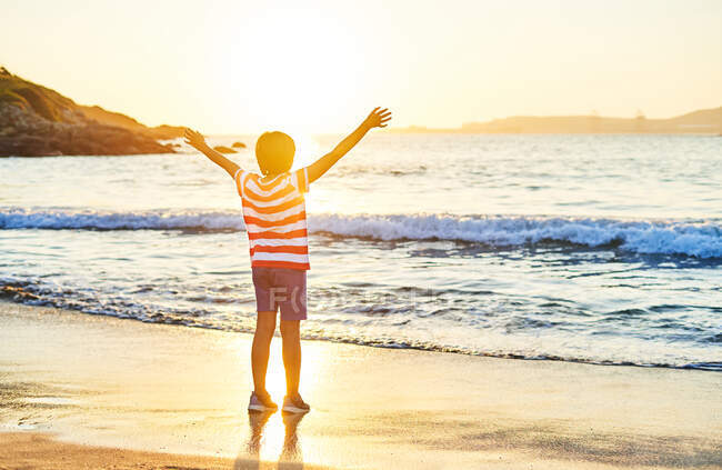 Вид ззаду на повну довжину невпізнаваного хлопчика, що стоїть з розтягнутими руками на мокрій піщаному березі, омитому хвилями блакитного моря на заході сонця — стокове фото