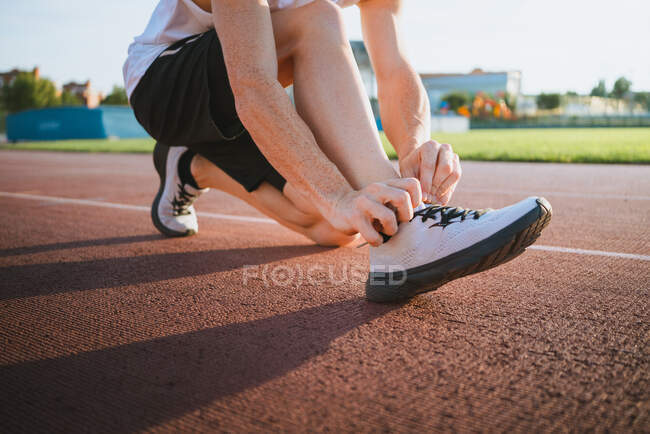 Crop unrecognizable male runner putting on modern sneaker while squatting on track before workout in sunlight — Stock Photo