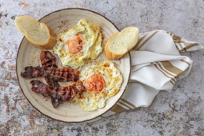 Overhead view of tasty sunny side up eggs with fried bacon strips and wheat bread pieces on plate above towel — Stock Photo