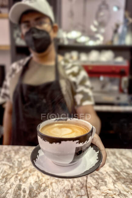 Homem étnico na máscara facial servindo xícara de café aromático com arte latte enquanto olha para a câmera na cafetaria — Fotografia de Stock