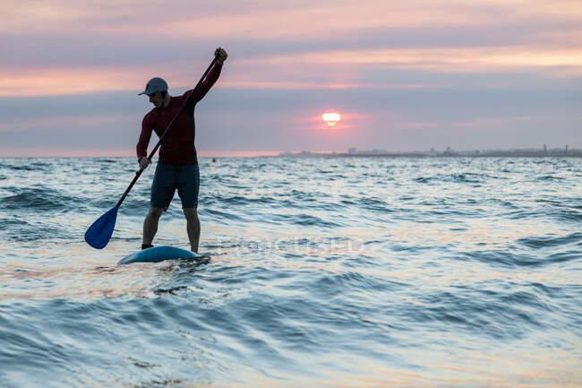 Мужчина-серфер в гидрокостюме и шляпе на весло доске для серфинга на берегу моря во время заката — стоковое фото