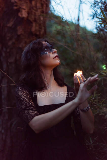 Таємнича відьма в чорному одязі і зі свічкою, що світиться в похмурому лісі і дивиться вниз — стокове фото