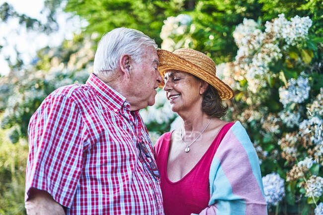 Sanftes Seniorenpaar streichelt zärtlich die Stirn, während es in der Natur neben blühenden Sträuchern steht — Stockfoto