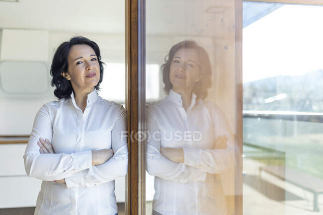 Mature confiant femme debout regardant la caméra avec les bras croisés appuyé sur la porte en verre regardant la caméra le matin à la maison — Photo de stock