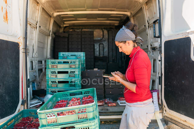 Jardinero femenino concentrado en bandana que navega por el teléfono celular mientras trabaja en la granja y mira la pantalla - foto de stock