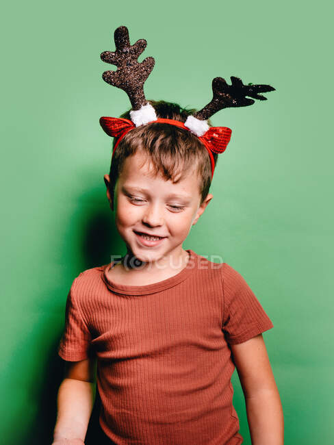 Улыбающийся мальчик с оленьими рогами, стоящий на зеленом фоне и смотрящий вниз — стоковое фото