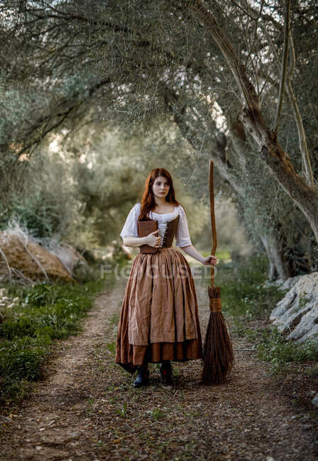 Grave strega in abito in piedi con libro magico di incantesimi e scopa su strada nel bosco e guardando altrove — Foto stock