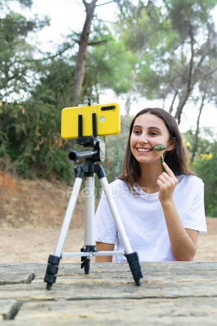 Веселая девушка подросток-блогер массирует щеку нефритовым роликом во время записи видео на мобильный телефон в парке — стоковое фото