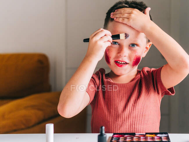 Charming child with makeup applicator touching head while looking at camera at table with eyeshadow palette — Stock Photo