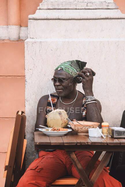 Прохолодний дорослий афроамериканець у сонячних окулярах і браслетах сидить за столом у міській кафетерії з кокосовим напоєм. — стокове фото