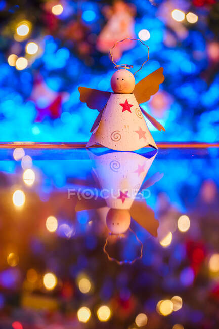 Handgemachtes engelförmiges Spielzeug auf Spiegeltisch gegen festlichen Weihnachtsbaum mit leuchtenden Girlanden gestellt — Stockfoto