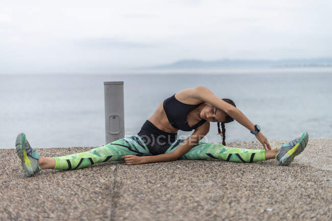 Fit female athlete sitting on embankment and doing side bend while stretching muscles and warming up during training — Stock Photo