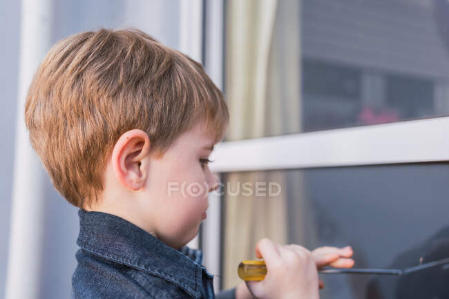 Vista lateral del niño enfocado aprendiendo a usar el destornillador mientras se refleja en la ventana durante el día - foto de stock