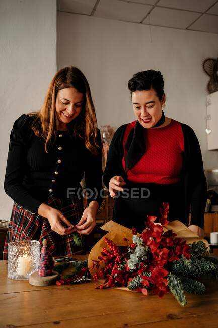 Веселі друзі-жінки стоять за столом зі свічками і роблять творчі різдвяні букети для святкування свята — стокове фото