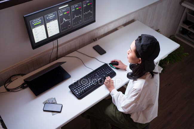 Високий кут зосереджує азіатську жінку, яка працює на комп'ютері з графіками, що показують динаміку змін в вартості криптовалют на зручному робочому місці. — стокове фото