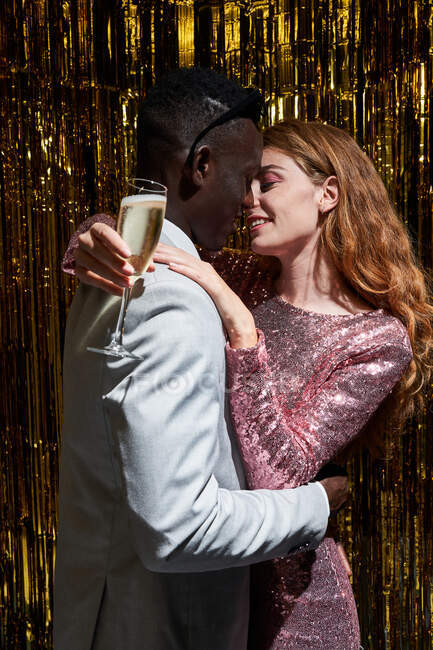 Elegante casal multiétnico com copo de champanhe abraçando e beijando cara durante a celebração de Ano Novo — Fotografia de Stock