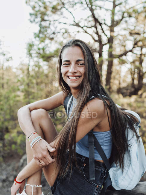 Smiling female teenager in trendy gumshoe and backpack touching forearm while looking at camera on fence in Tenerife Spain — Stock Photo
