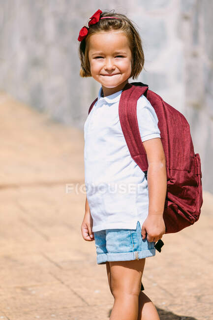 Vista lateral da criança em idade escolar com mochila no pavimento olhando para a câmera à luz do sol — Fotografia de Stock
