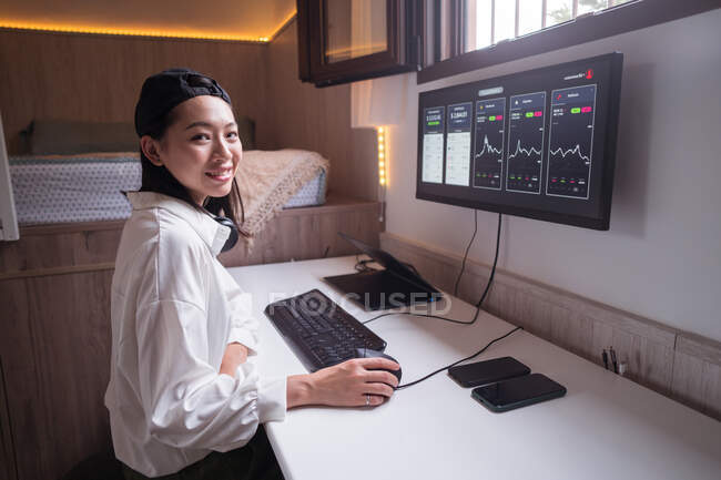 Seitenansicht einer fröhlichen Asiatin, die am Computer arbeitet, mit Diagrammen, die dynamische Wertveränderungen der Kryptowährung am bequemen Arbeitsplatz zeigen — Stockfoto