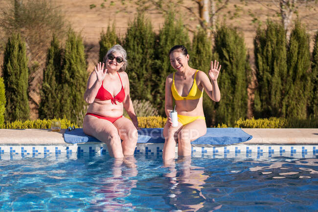 Old woman in sunglasses sitting near Asian daughter on poolside and waving hands to camera — Stock Photo