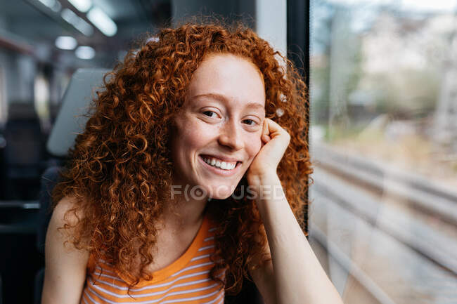 Cheerful young woman with curly red hair looking at camera during trip on train — Stock Photo