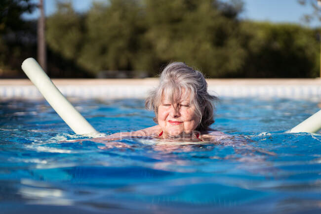 Веселая пожилая женщина с седыми волосами, плавающая в бассейне с аква-лапшой — стоковое фото