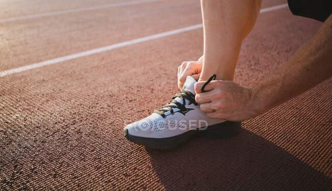 Crop unrecognizable male runner putting on modern sneaker while squatting on track before workout in sunlight — Stock Photo