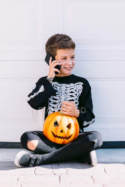Полное тело веселого мальчика в костюме черного скелета с раскрашенным лицом и вырезанной тыквой на Хэллоуин, разговаривающей по мобильному телефону, сидя возле белой стены на улице — стоковое фото