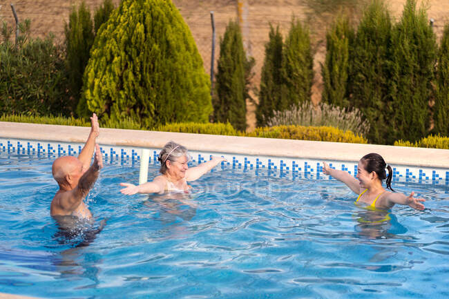 Усміхнена сім'я піднімає руки під час занять водною аеробікою у басейні з чистою блакитною водою — стокове фото