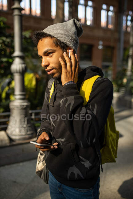 African American male hipster in street style clothes and wireless earphones listening to music while standing in city street and messaging on mobile phone — Stock Photo