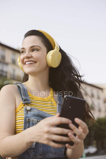De baixo feminino despreocupado em fones de ouvido sem fio navegando no smartphone na rua olhando para longe — Fotografia de Stock