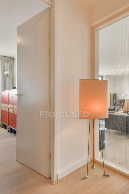 Porte ouverte entre armoires et lampe sur parquet dans un espace de travail lumineux avec mur de verre contre canapé — Photo de stock