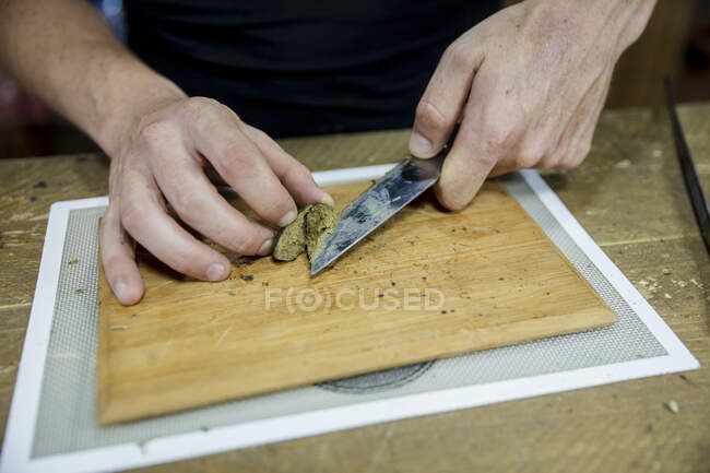 Crop unrecognizable male with knife cutting dried cannabis plant piece on wooden board in workspace — Stock Photo