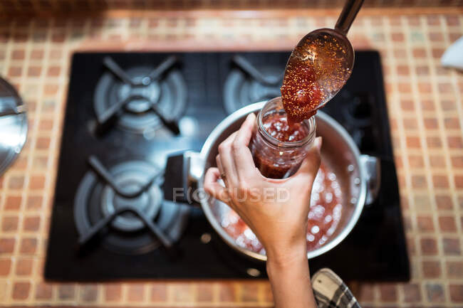 High angle of crop anonymous person pouring delicious fig confiture into jar above gas stove at home — Stock Photo