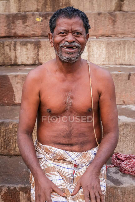 INDIA, VARANASI - 27 DE NOVIEMBRE DE 2015: Varón desdentado de mediana edad, sin camisa, de pelo gris y con ropa casual, de pie cerca de un edificio en mal estado y mirando a la cámara - foto de stock