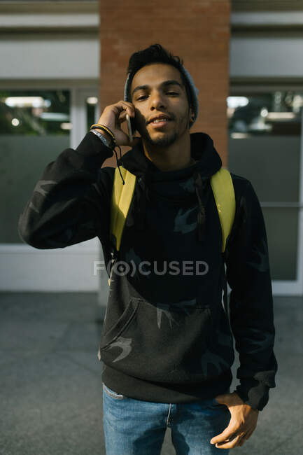 Handsome African American male hipster in hat and hoodie smiling and looking at camera while standing speaking on smartphone in urban street — Stock Photo