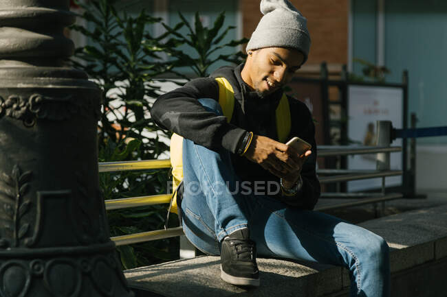 Contenido Hipster masculino afroamericano con mochila sentada en la frontera de piedra en la calle y mensajería en el teléfono móvil - foto de stock